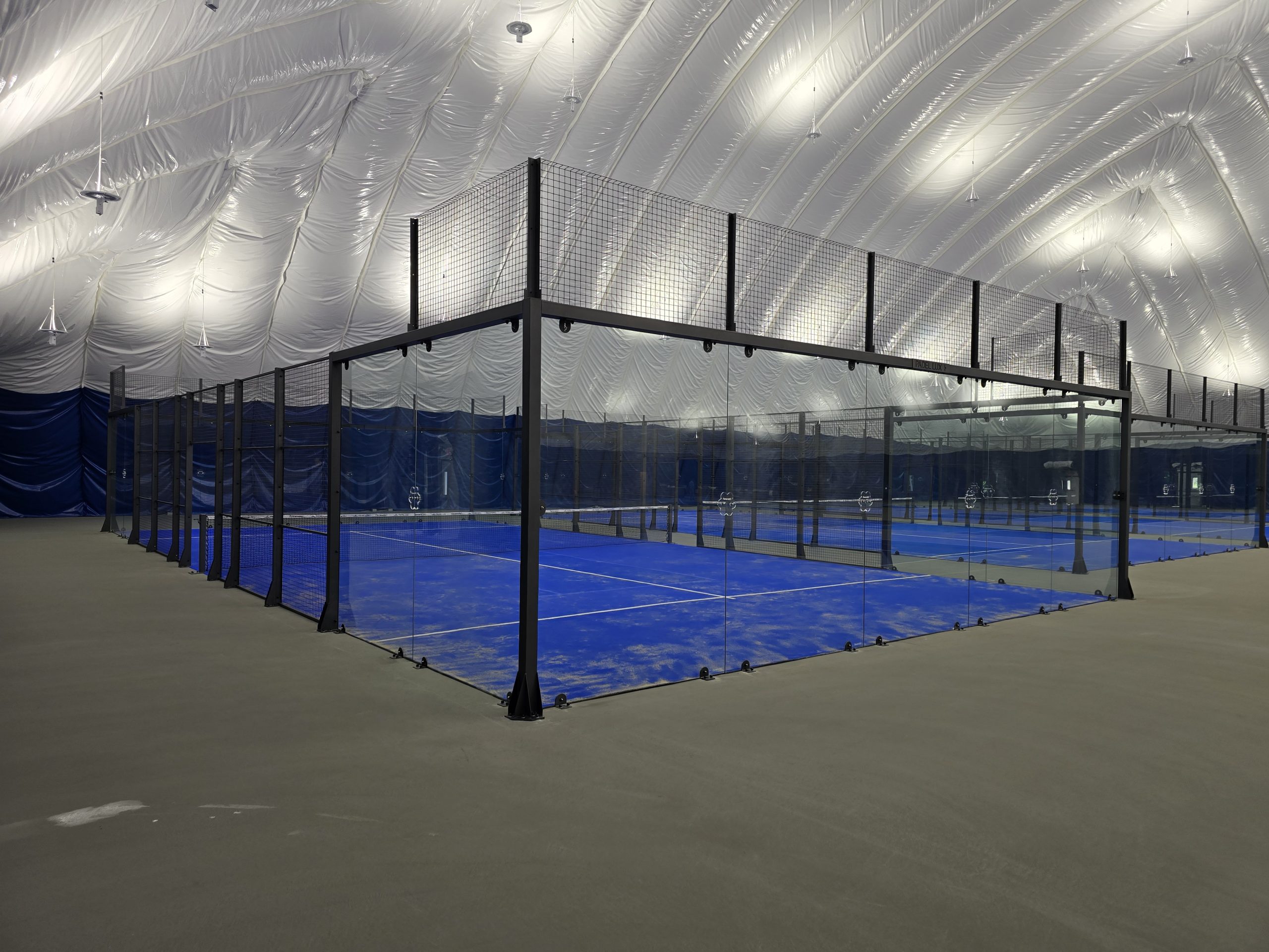 20250806_101450 Indoor pickleball courts with a blue playing surface, enclosed by black fencing and surrounded by a high ceiling.