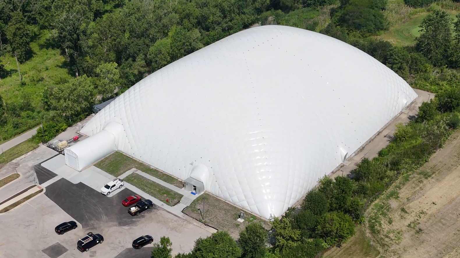 Screenshot 2026-03-10 105045 Aerial view of a large, white domed structure surrounded by greenery and parked vehicles, indicating a recreational facility.