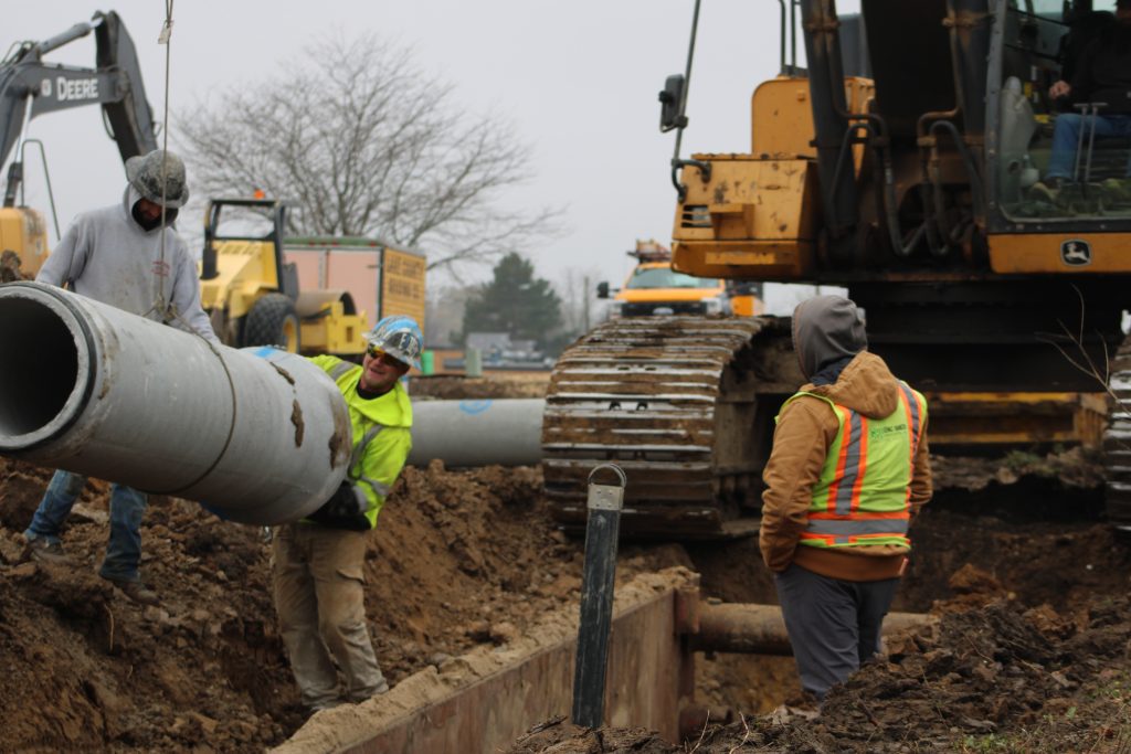 Construction workers guide a large concrete pipe into a trench using heavy machinery at an infrastructure project site