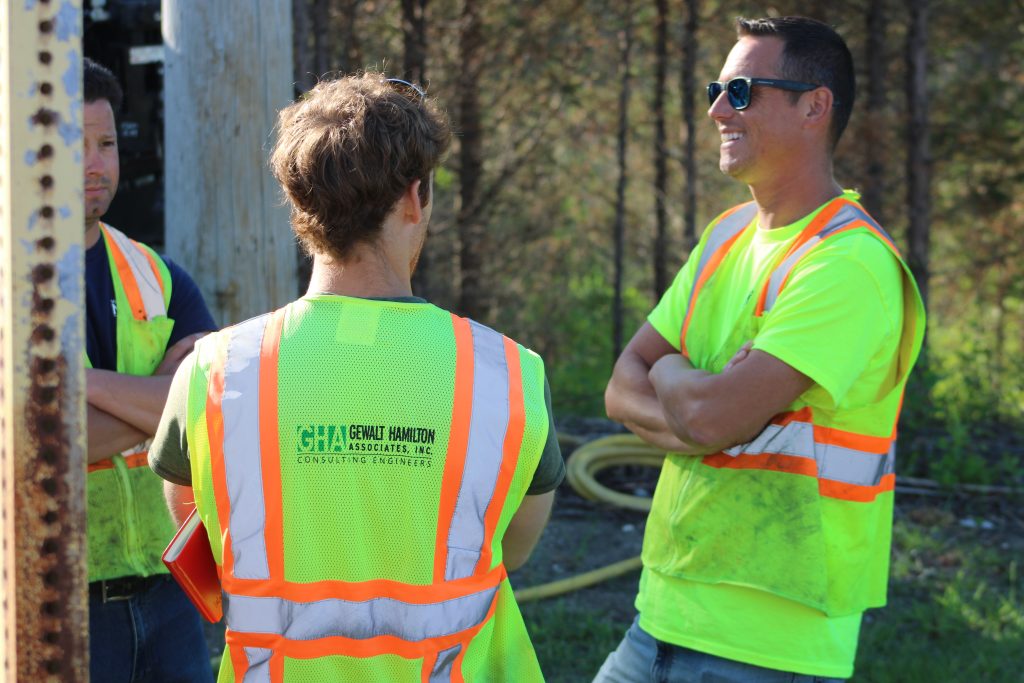 Three engineers wearing high-visibility safety vests with the Gewalt Hamilton Associates logo talk at a job site